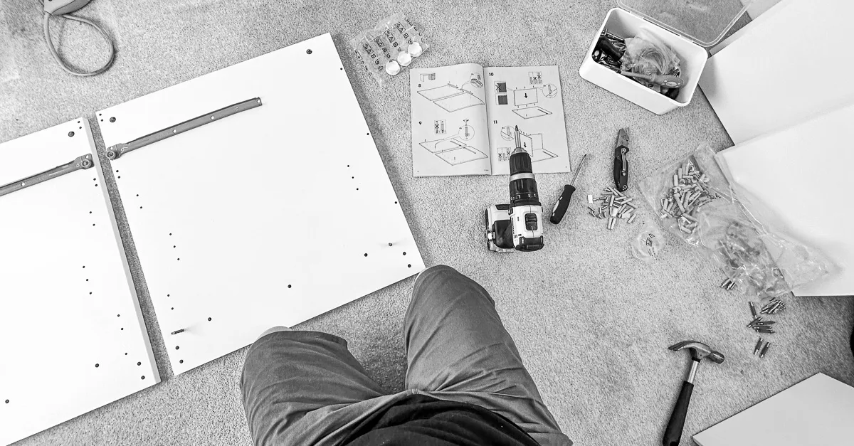 A black and white of ikea desk parts and fittings layed out on the carpet before me ready for assembly