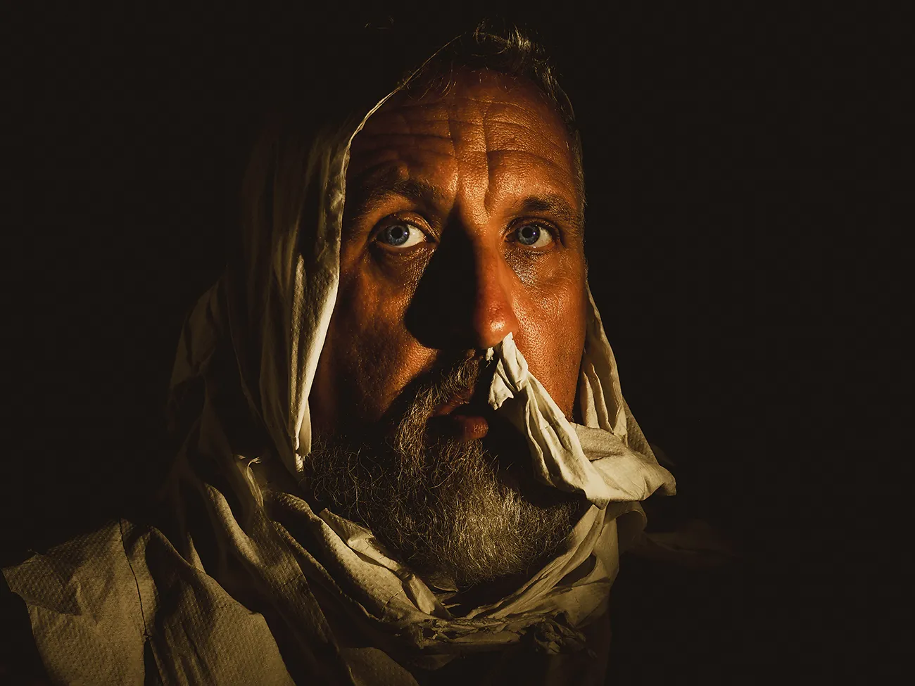 Stuart against a dark backdrop, lit using a small amount of light, he is swaddled in clothes made of bogroll channeling a lawrence of arabia look