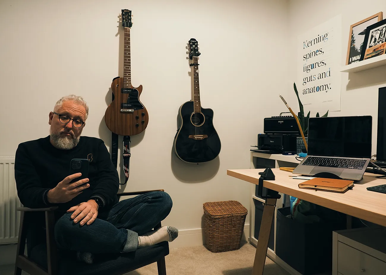 man sits alone in a chair with his phone, he&#x27;s in his home office surrounded by desk, laptop &#x26; guitars