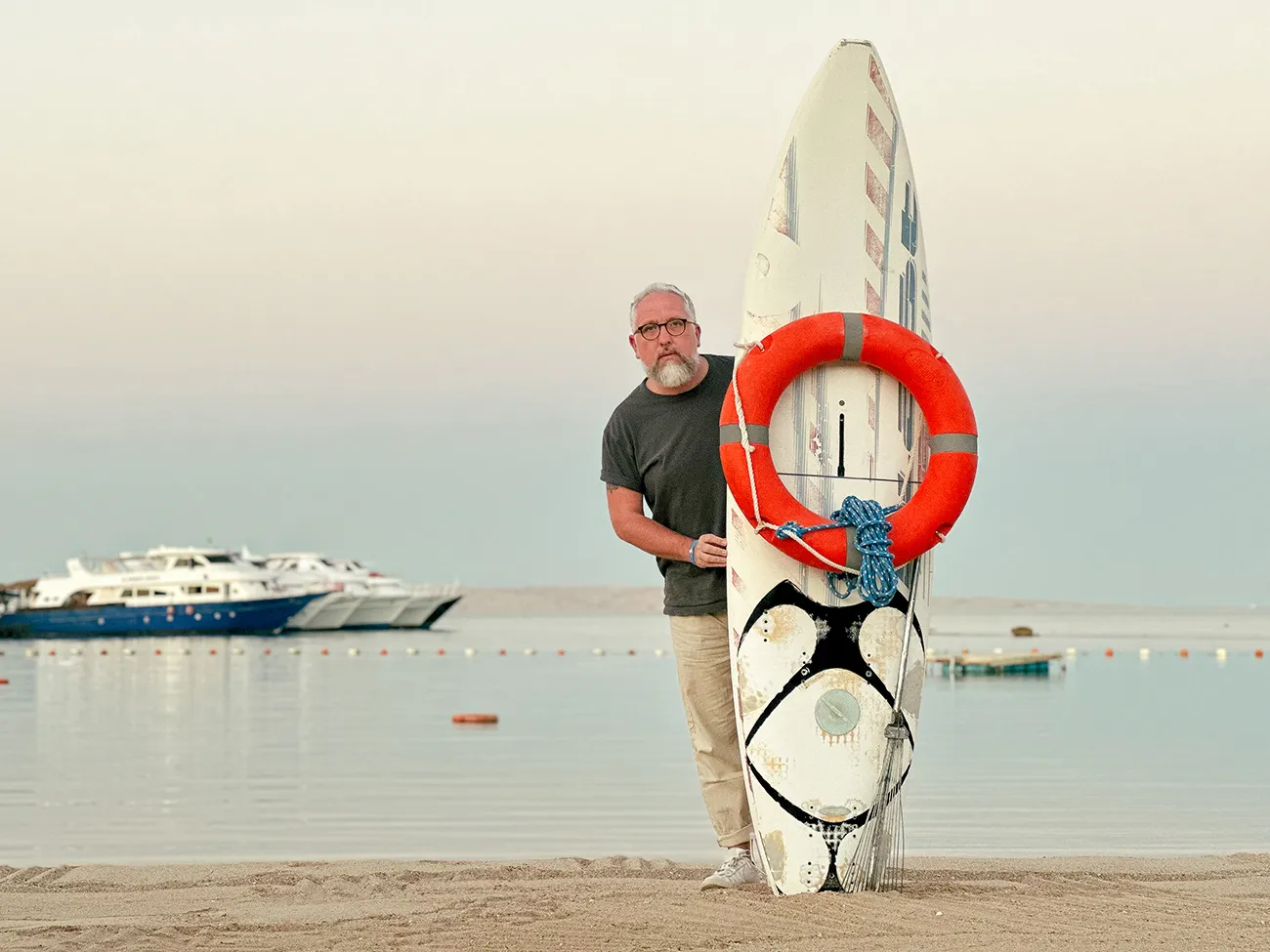 A surf board stands upright on the short of a beach, Stu peers out from behind it!