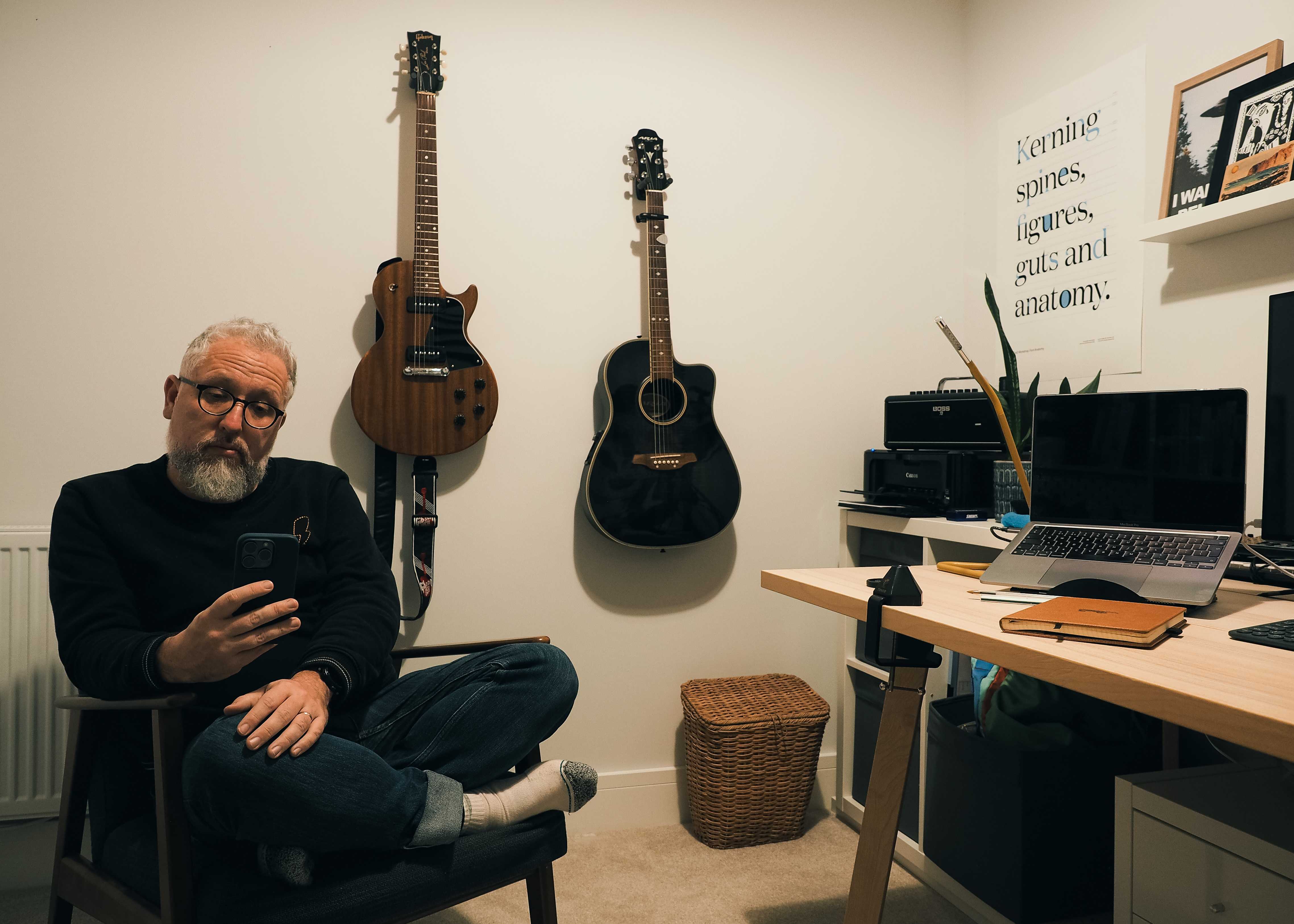 man sits alone in a chair with his phone, he's in his home office surrounded by desk, laptop & guitars